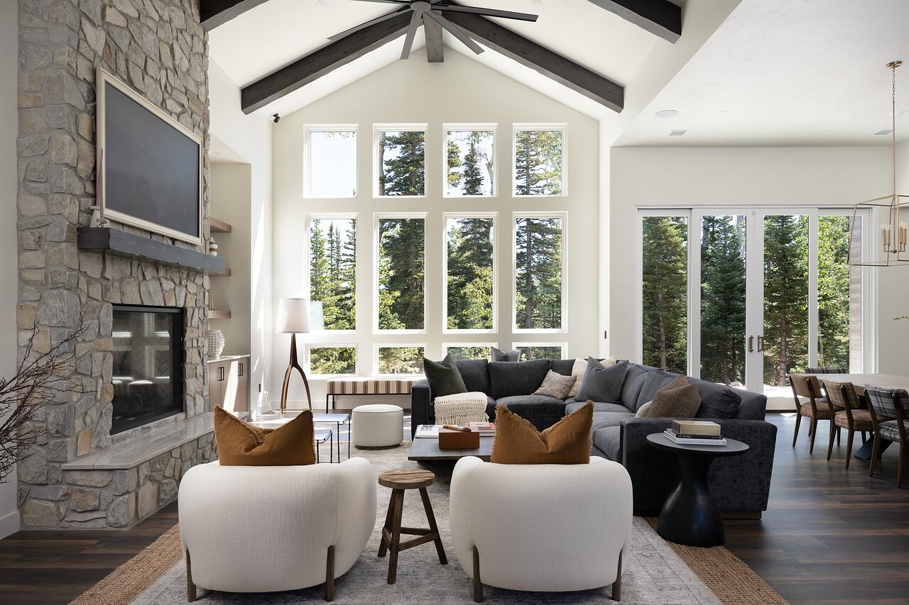 Vaulted great room with a floor-to-ceiling stone fireplace, black beams, cream curved chairs, a charcoal sectional, and tall windows onto the pines
