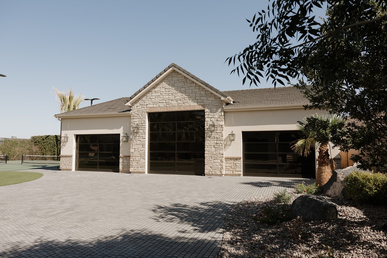 Three-bay garage seen straight on: a ledgestone center gable, cream stucco wings, full-glass doors, lantern sconces, and a paver forecourt